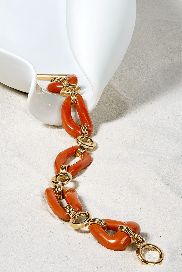Schmuck-Moodfotografie eines Gliederarmbands in Korallenrot und Gelbgold an einer geschwungenen weißen Porzellanschale auf hellem Sand